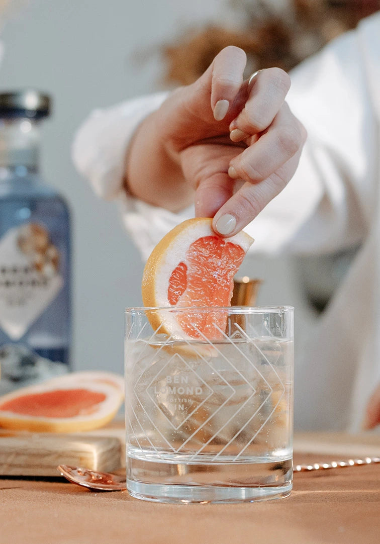 Hand placing a grapefruit slice into a glass with a bottle of gin in the background
