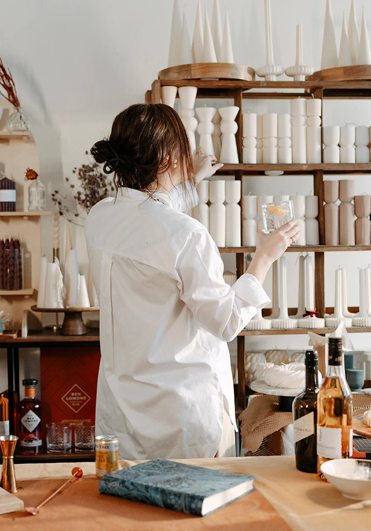 Person in a white shirt standing in a store with shelves of products and a table with bottles and a book.