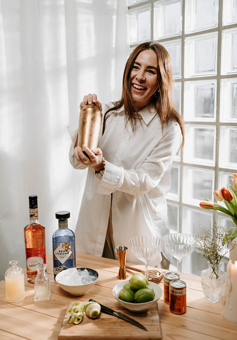 Woman making a cocktail in a kitchen with various ingredients and bottles on the table.