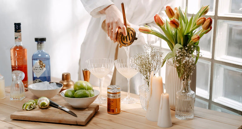 Person preparing a drink at a table with bottles, fruits, and a vase of flowers.