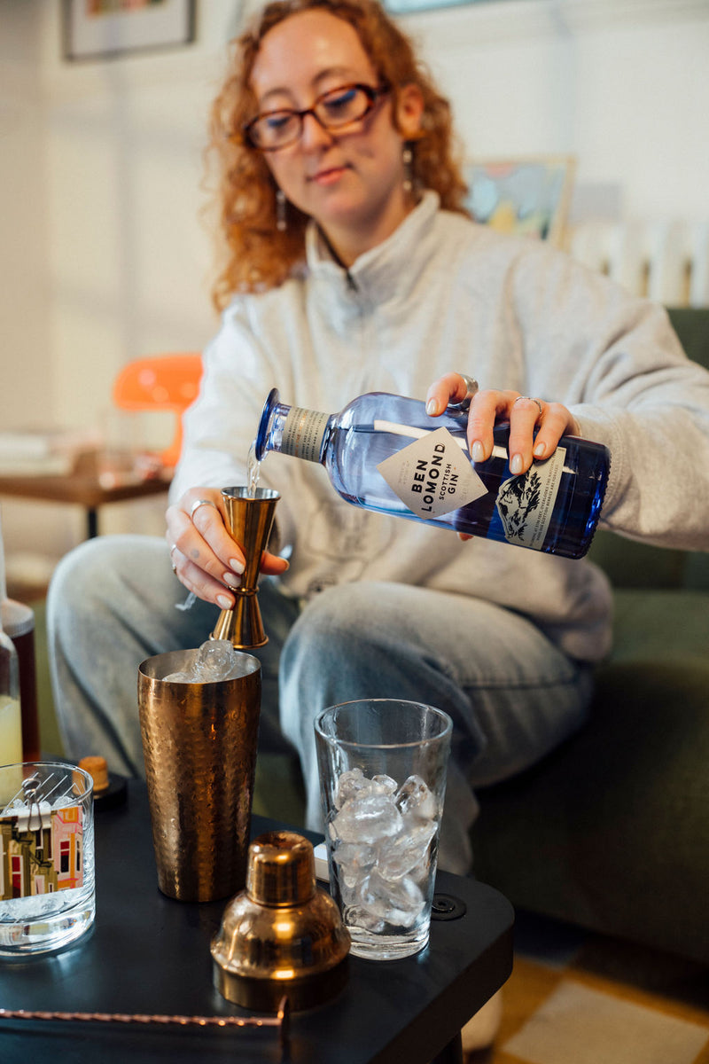 Person pouring a drink from a blue bottle into a glass with ice on a table.
