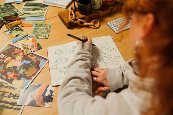 Person drawing in a notebook with various art supplies on a table