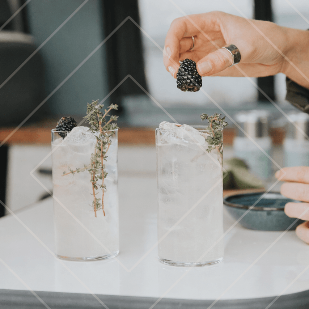 Person garnishing a cocktail with a blackberry next to two glasses filled with a white drink and garnishes.