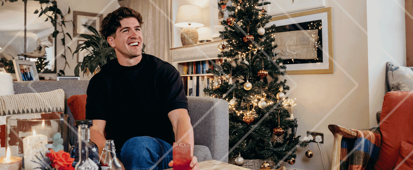 Man sitting in a cozy living room with a Christmas tree, holding a drink.