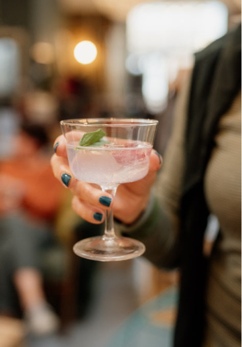 Hand holding a pink cocktail with a leaf garnish in a blurred indoor setting