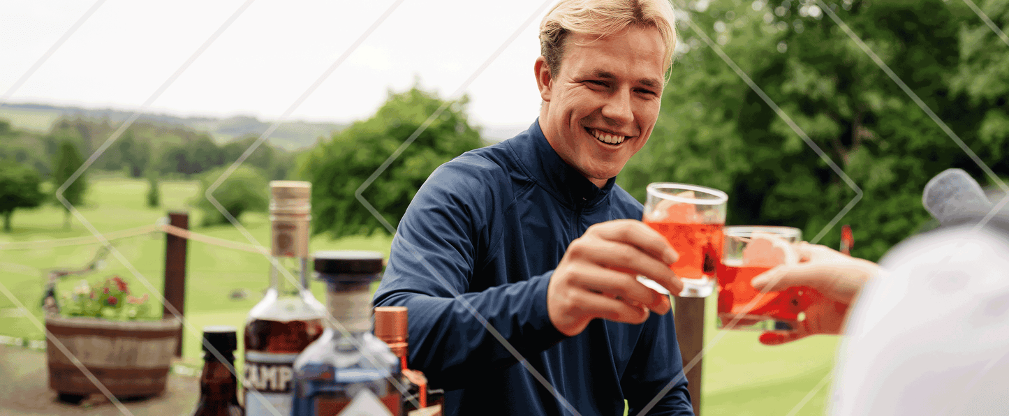 Man holding a glass of red liquid with another person outdoors, surrounded by bottles on a table.