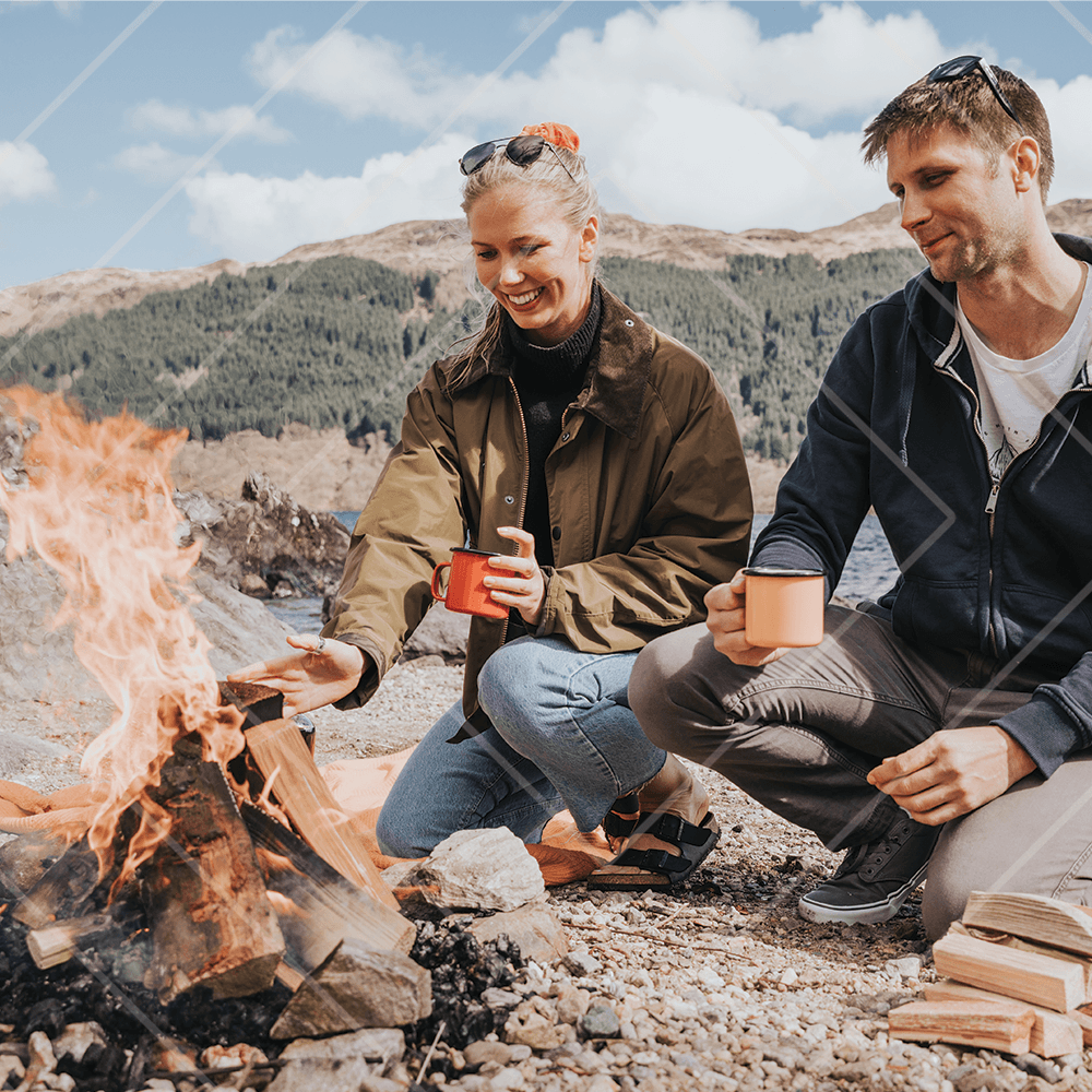 Two people sitting by a lakeside campfire, smiling and holding enamel mugs, with mountains and trees in the background on a bright day.