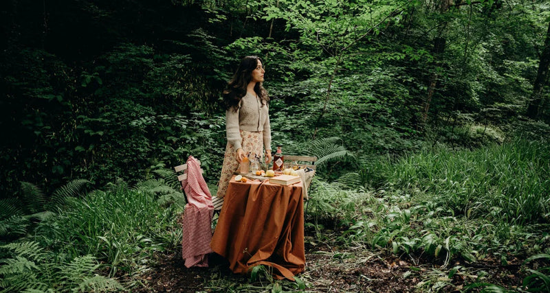 Woman standing in a forest with a table set up, surrounded by greenery.