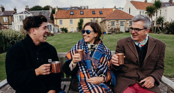 Three people sitting outdoors, holding mugs, with a scenic background of houses and greenery.