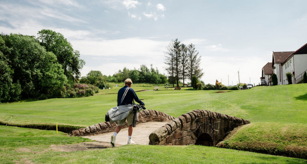 Golfer walking across a bridge on a golf course with trees and a building in the background.