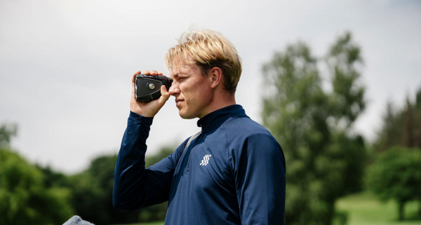 Man in a blue jacket using a golf rangefinder on a golf course