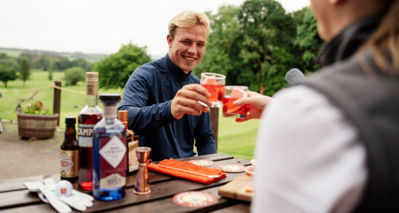 Two people enjoying drinks outdoors at a picnic table with food and drinks.