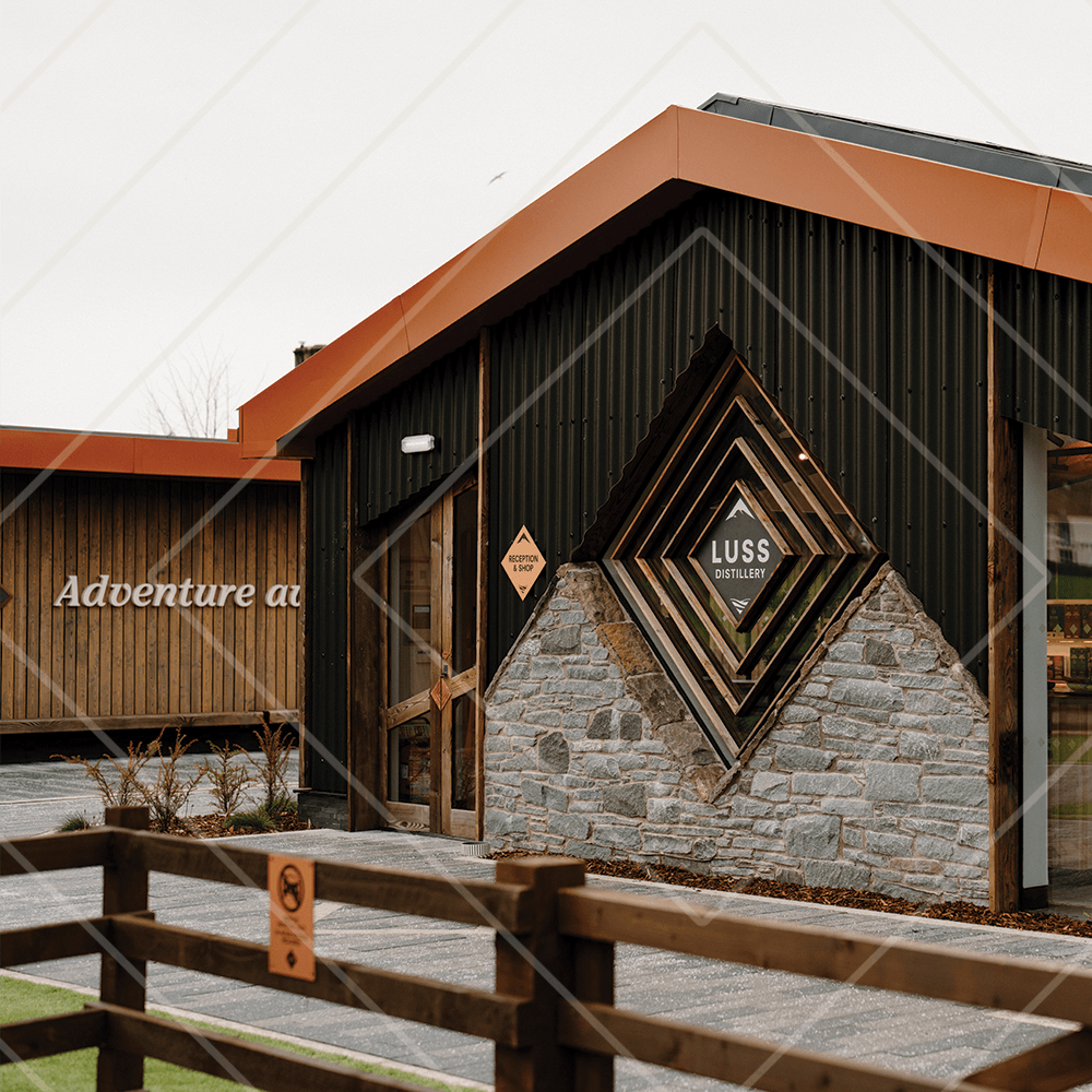 Exterior of Luss Distillery with modern wood, stone, and black metal design, featuring a geometric window and signage on an overcast day.
