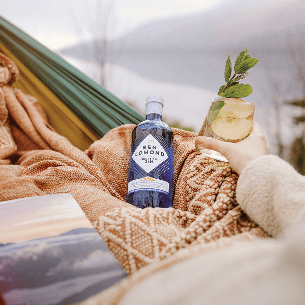 Person holding a bottle of Ben Lomond Gin and a cocktail in a hammock outdoors.