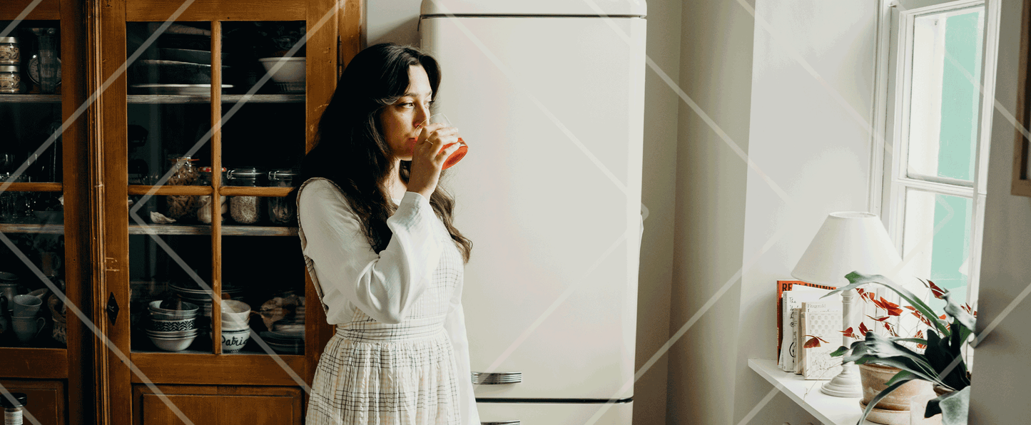 Woman in a white dress standing in a kitchen, holding a cup.