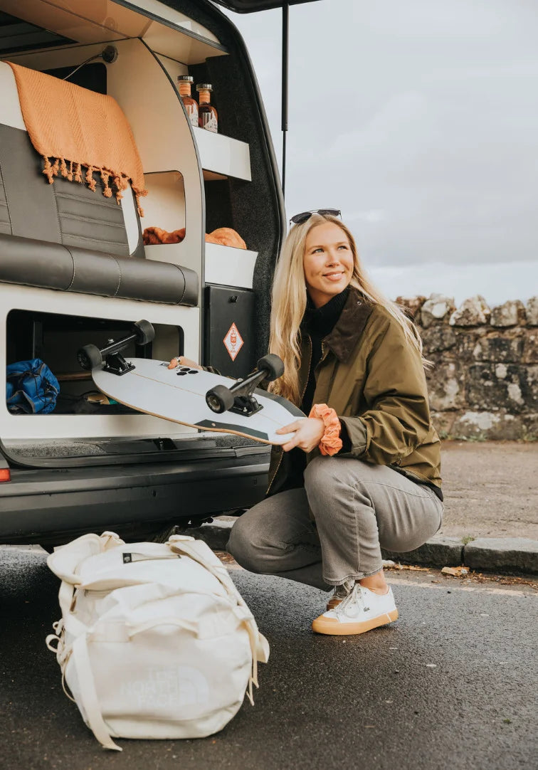 Woman holding a snowboard next to a van with camping gear, outdoors.