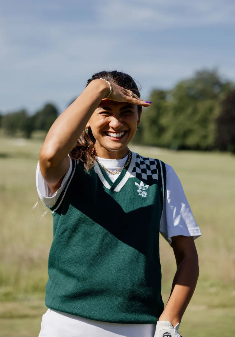 Person on a golf course wearing a green vest and white shirt, with a blurred background of trees and sky.