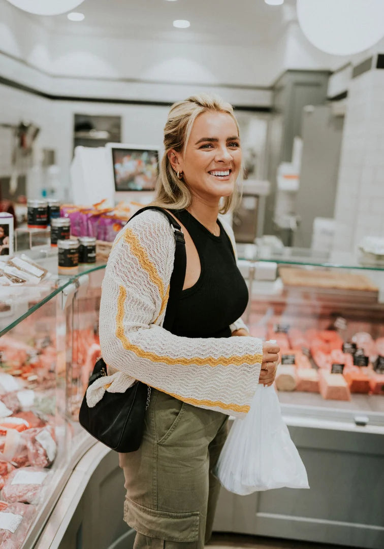 Woman standing in a grocery store holding a bag, wearing a black top and beige cardigan.
