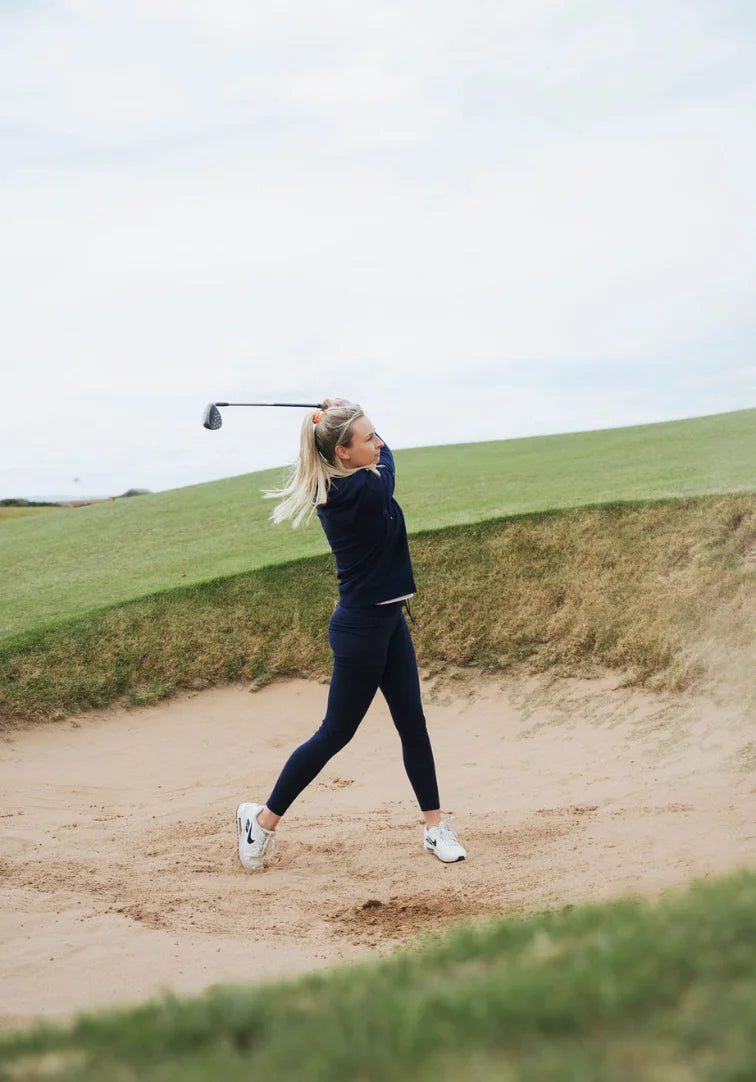 Woman playing golf in a sand trap on a grassy course with a clear sky.