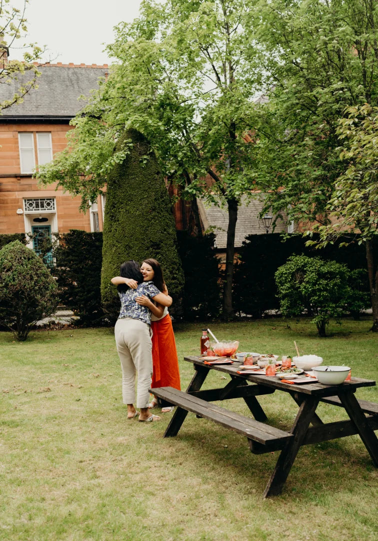 Two women hugging in a garden with a picnic table and food.