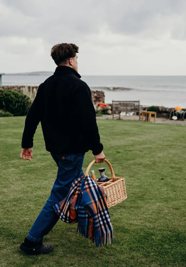 Man walking on grass with a picnic basket and plaid blanket, looking at the sea.