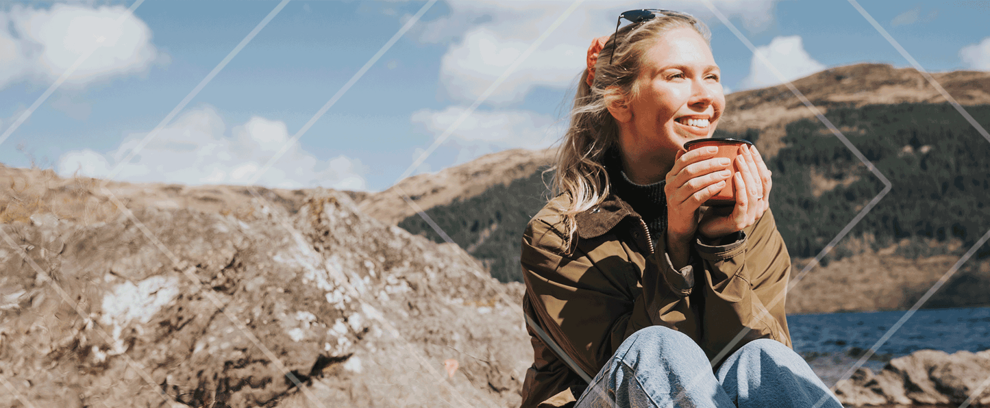 Woman sitting on a rocky outcrop holding a mug with mountains and sky in the background