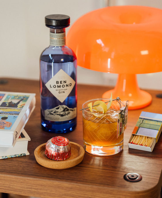 Bottle of Ben Lomond gin with a cocktail on a wooden table, featuring an orange lamp in the background.