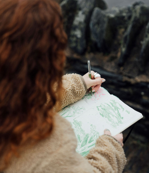 Person holding a drawing pad with green artwork and a pen, wearing a brown jacket.