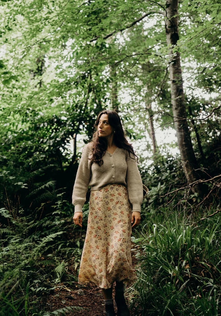 Woman standing in a forest wearing a beige sweater and floral skirt.