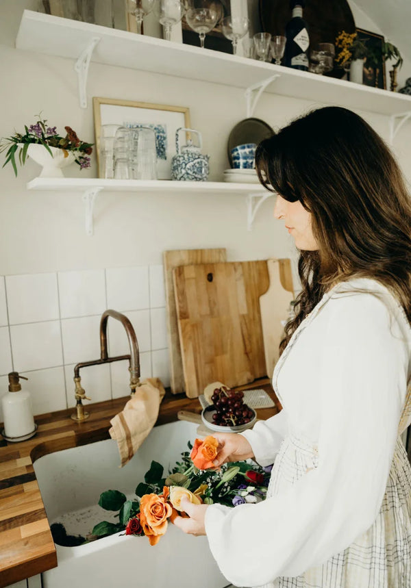 Woman arranging flowers in a kitchen with shelves and a sink in the background