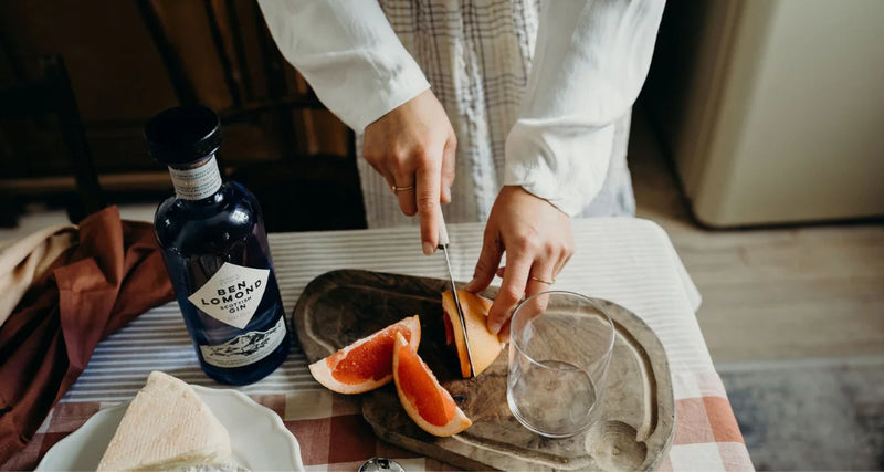 Person cutting a grapefruit on a wooden tray with a bottle of gin and a glass in the background.