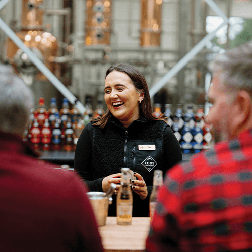 Woman in a black vest with a name tag standing behind the distillery bar, smiling at two people in front of her.