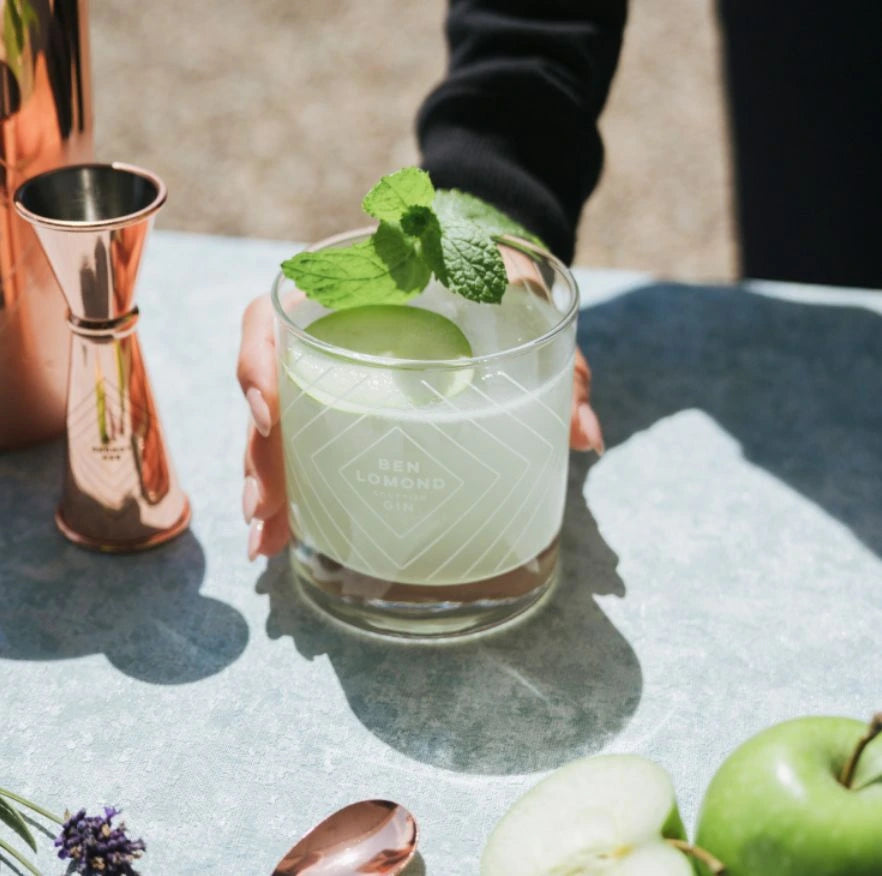 Cocktail glass with green drink and mint leaves held by a person, with a copper shaker and apples on a table.